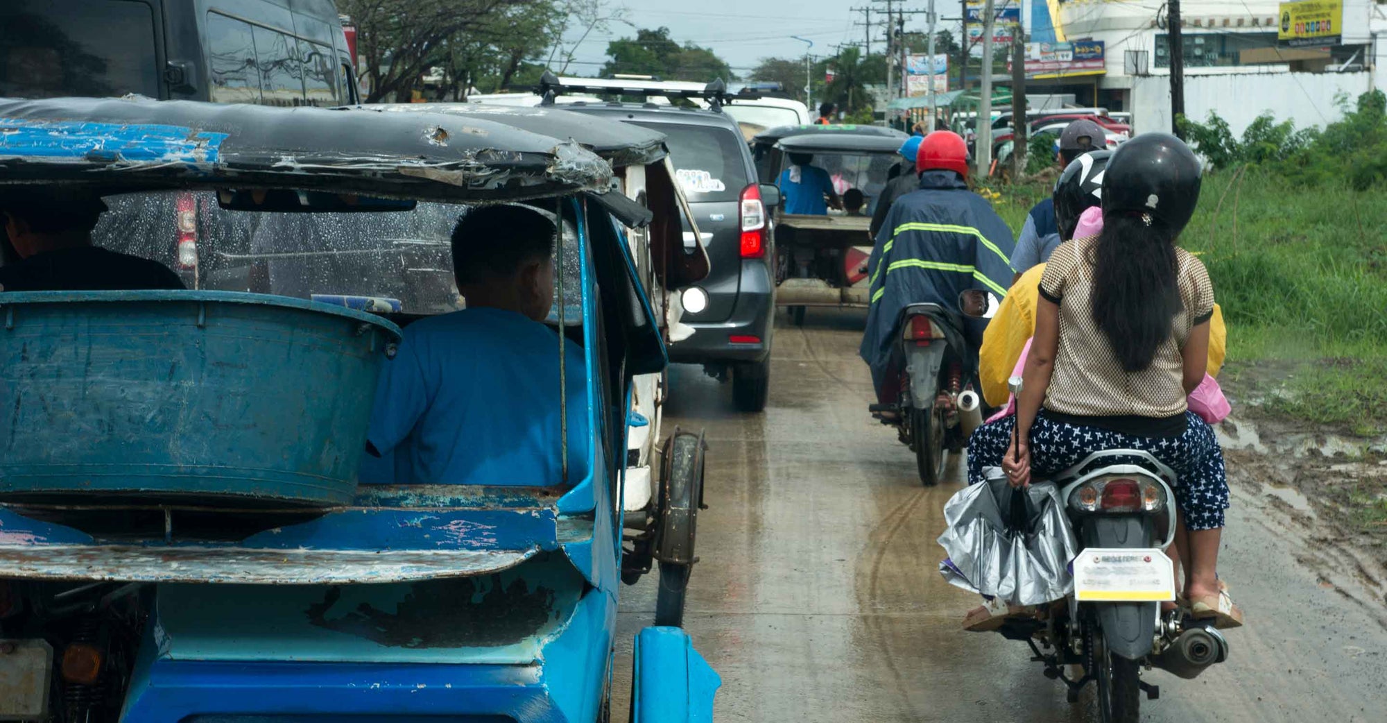 Filipino/a drivers on motorcycles and in cars on a wet, congested road in what is ostensibly the Philippines.