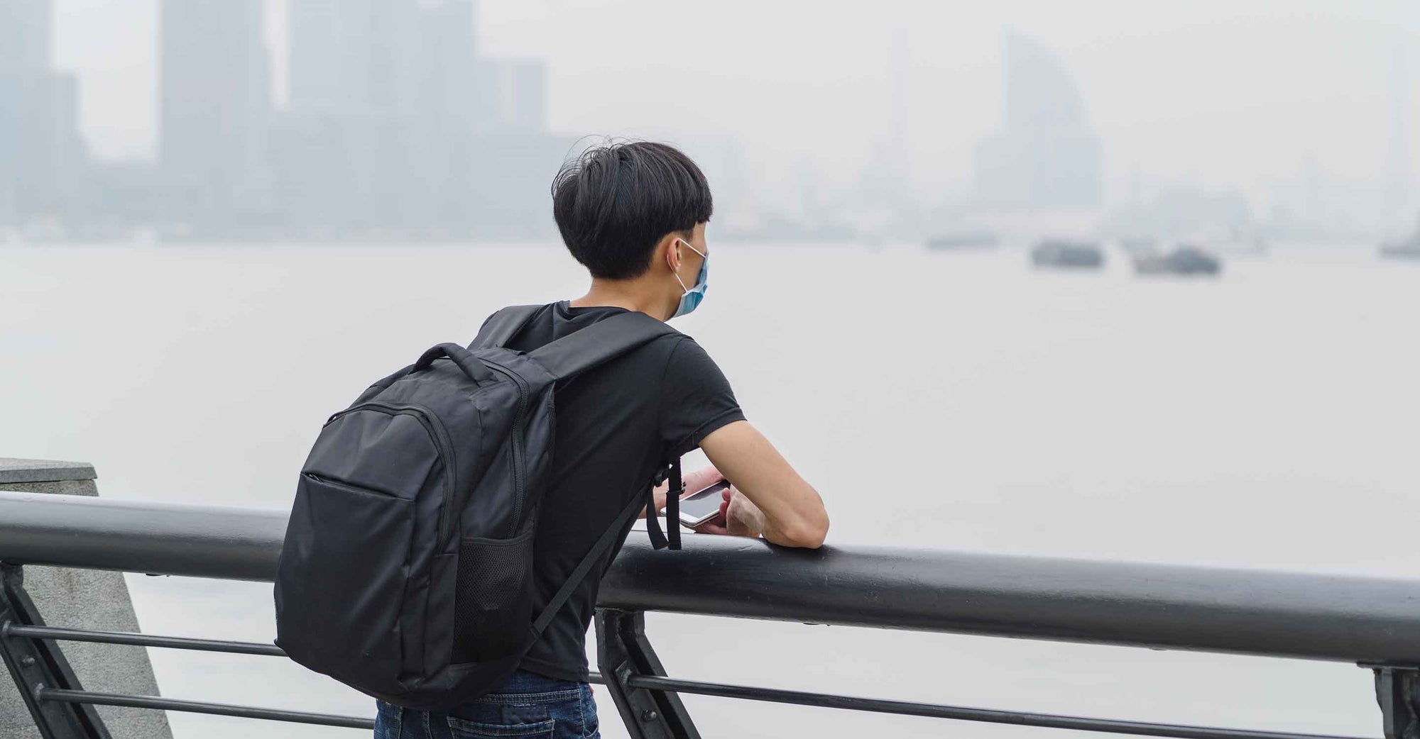 A young man looking out across the harbor of a major city, its skyline in the distance, that appears shrouded in a polluted haze.